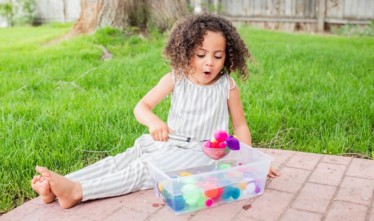 Young child using a cup to scoop up colorful pom poms